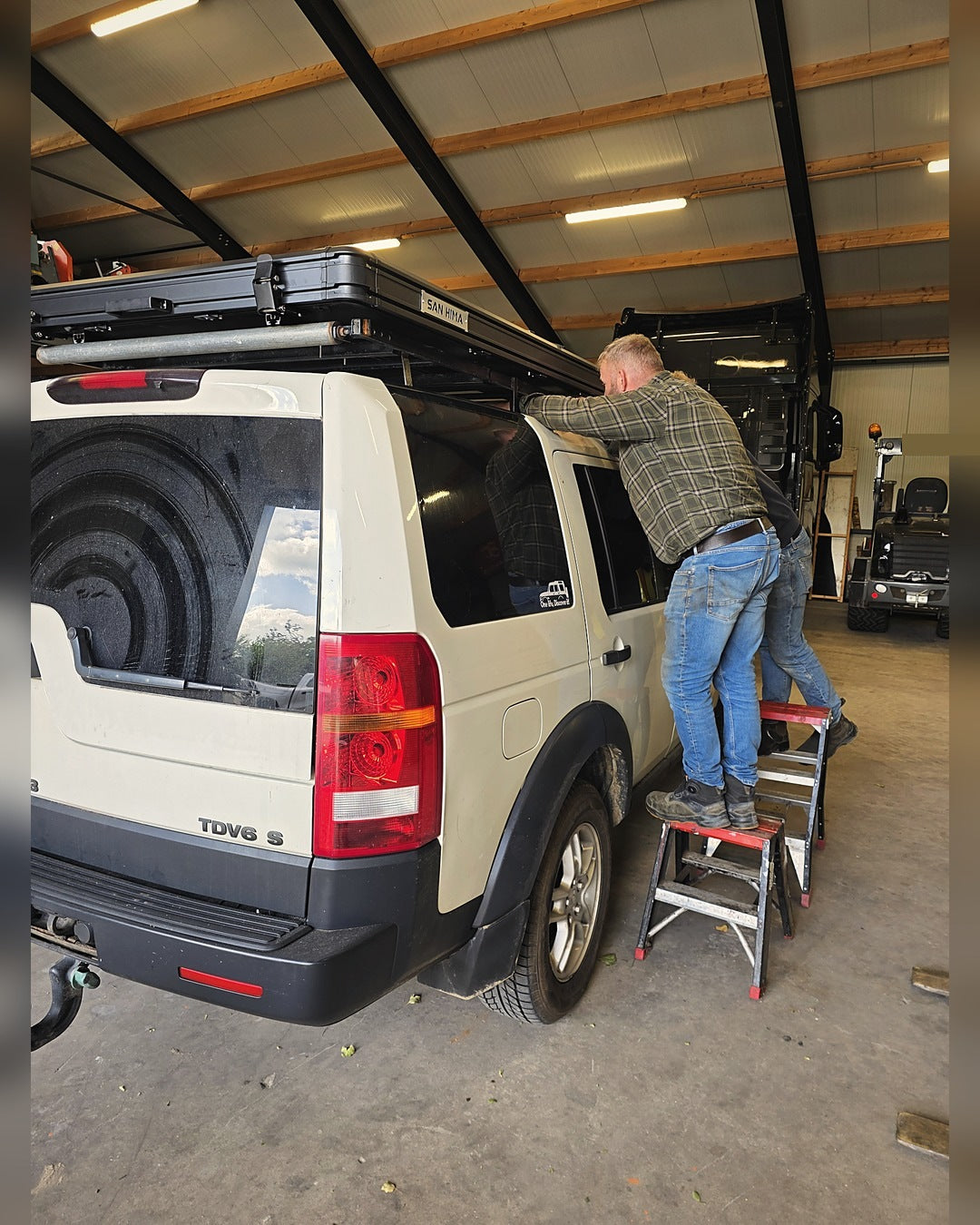 A hardshell rooftop tent being placed on the roof rack of a Land Rover Discovery in the workshop